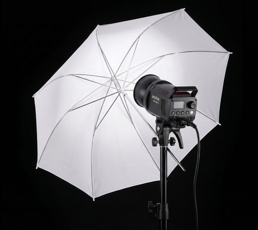 Le flash de studio Godox monté sur son pied, avec son grand parapluie
                    blanc ouvert qui diffuse la lumière.
